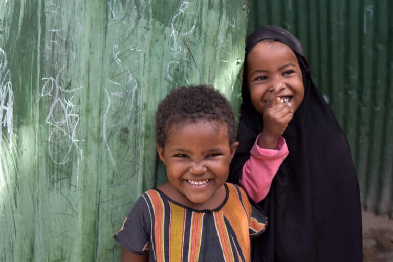 To jenter som står ved en grønn vegg mens de ler og smiler ved siden av hverandre.