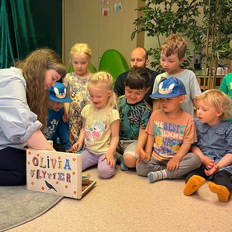Forfatter Agnes Thorstvedt, med mørkt langt hår og lyseblå skjorte, sitter på gulvet og leser for barn i Espira-barnehagen. Foto: Espira