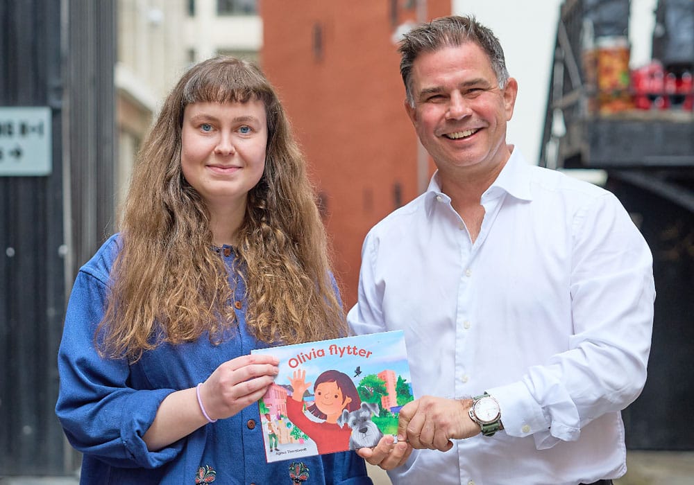 Forfatter Agnes Thorstvedt til venstre, sammen med Karsten Onsrud, administrerende direktør i Aktiv Eiendomsmegling, holder barneboka Agnes flytter mellom seg. Foto. Espira