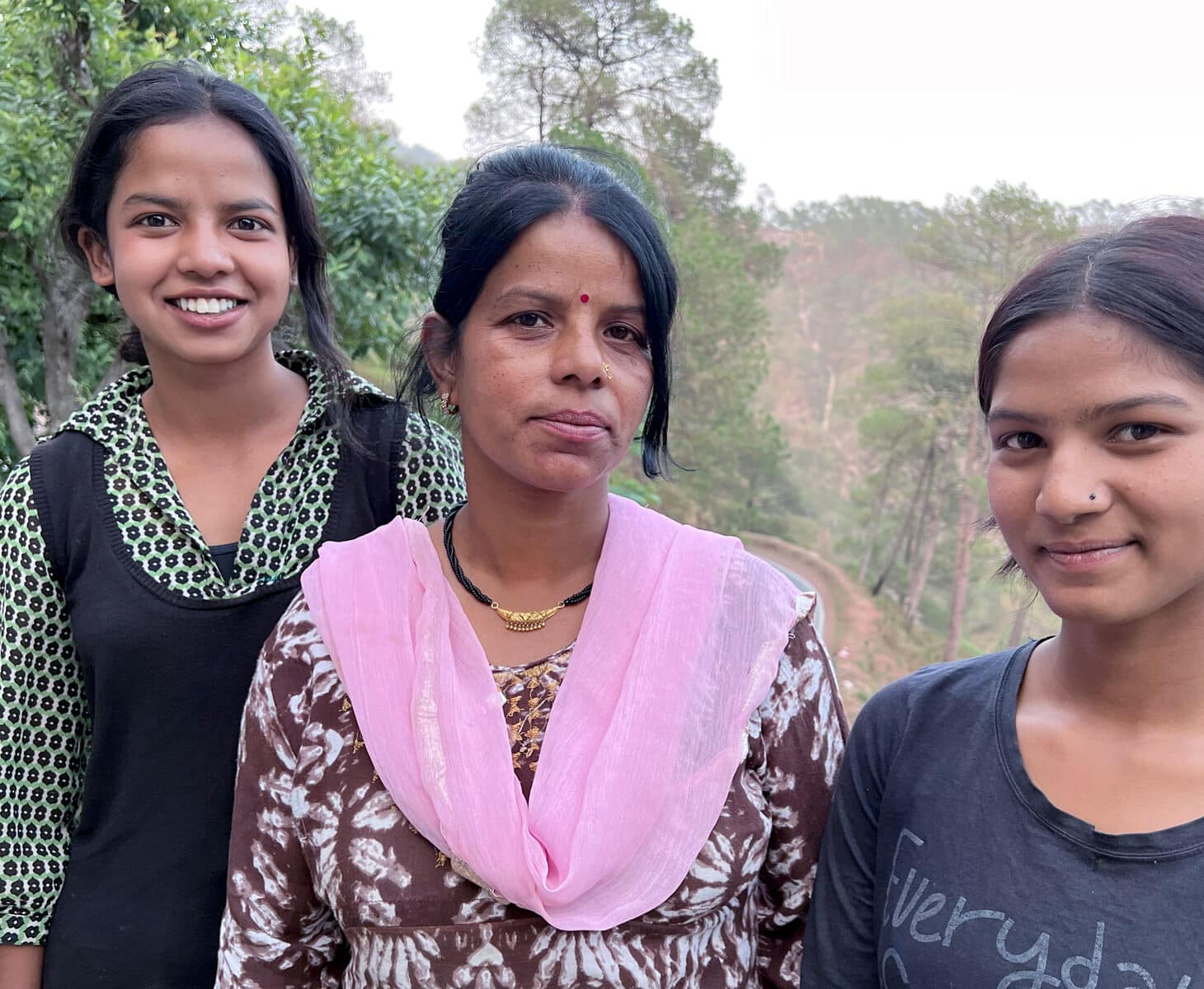 Manju Devi, med håret i hestehale, brun og hvit mønstret kjole og rosa skjerf, står sammen med døtrene. Den ene har svart veste og grønn mønstret skjorte, den andre blå t-skjorte.Foto: Pearl Sandhu