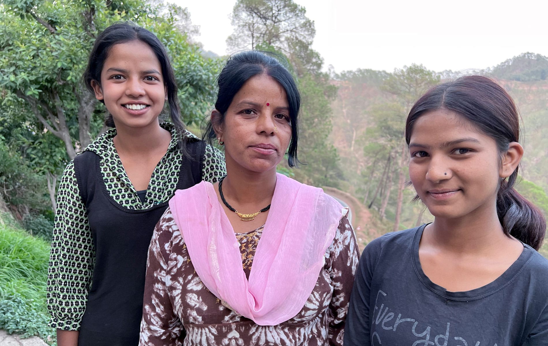Manju Devi, med håret i hestehale, brun og hvit mønstret kjole og rosa skjerf, står sammen med døtrene. Den ene har svart veste og grønn mønstret skjorte, den andre blå t-skjorte.Foto: Pearl Sandhu