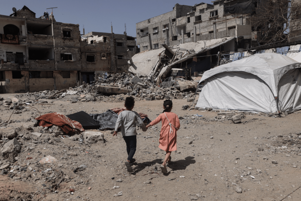 To barn holder hverandre i hendene og løper sammen mellom ruinerte bygninger i Rafah 2024.