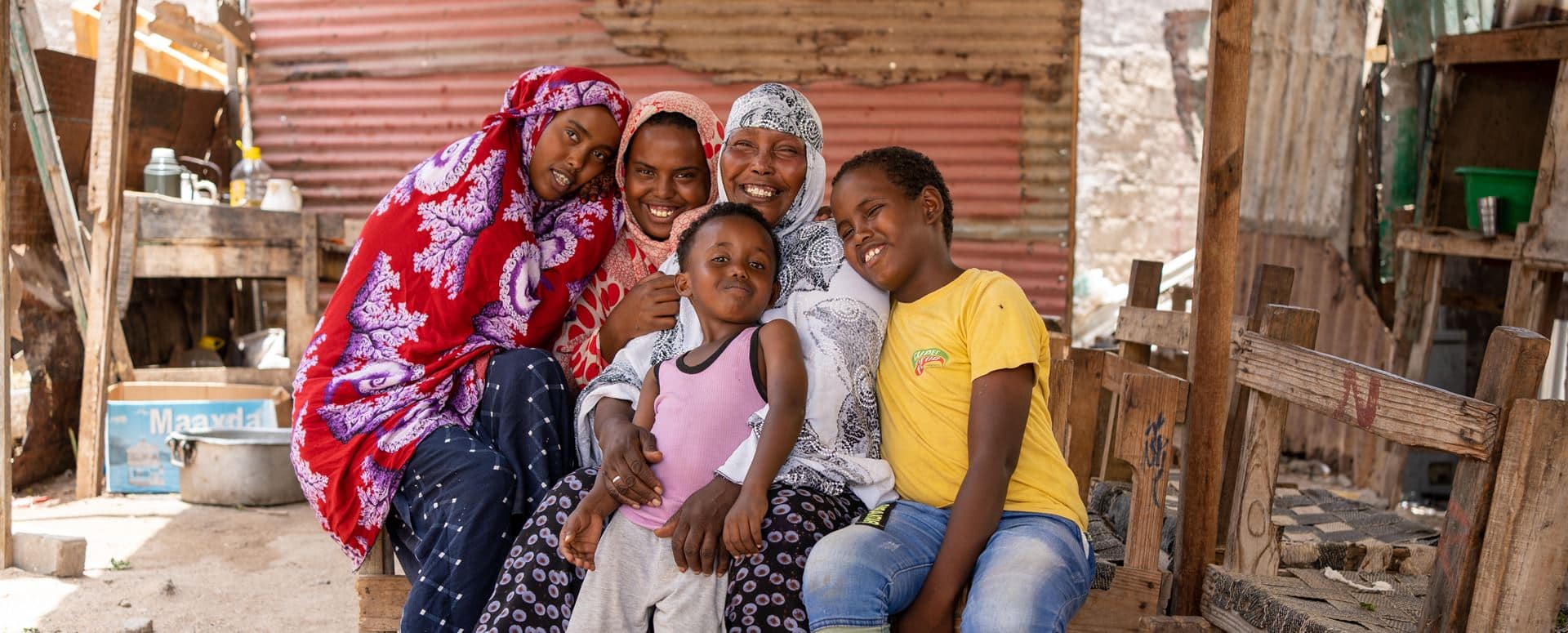 Hodan, med hvit mønstret hijab, sitter utenfor huset sammen med de fire barna. Jentene har fargerike hijaber, den minste gutten rosa t-skjorte og beige bukse, den eldste gutten gul ts-skjorte og jeans. Foto: Lydia Mantler