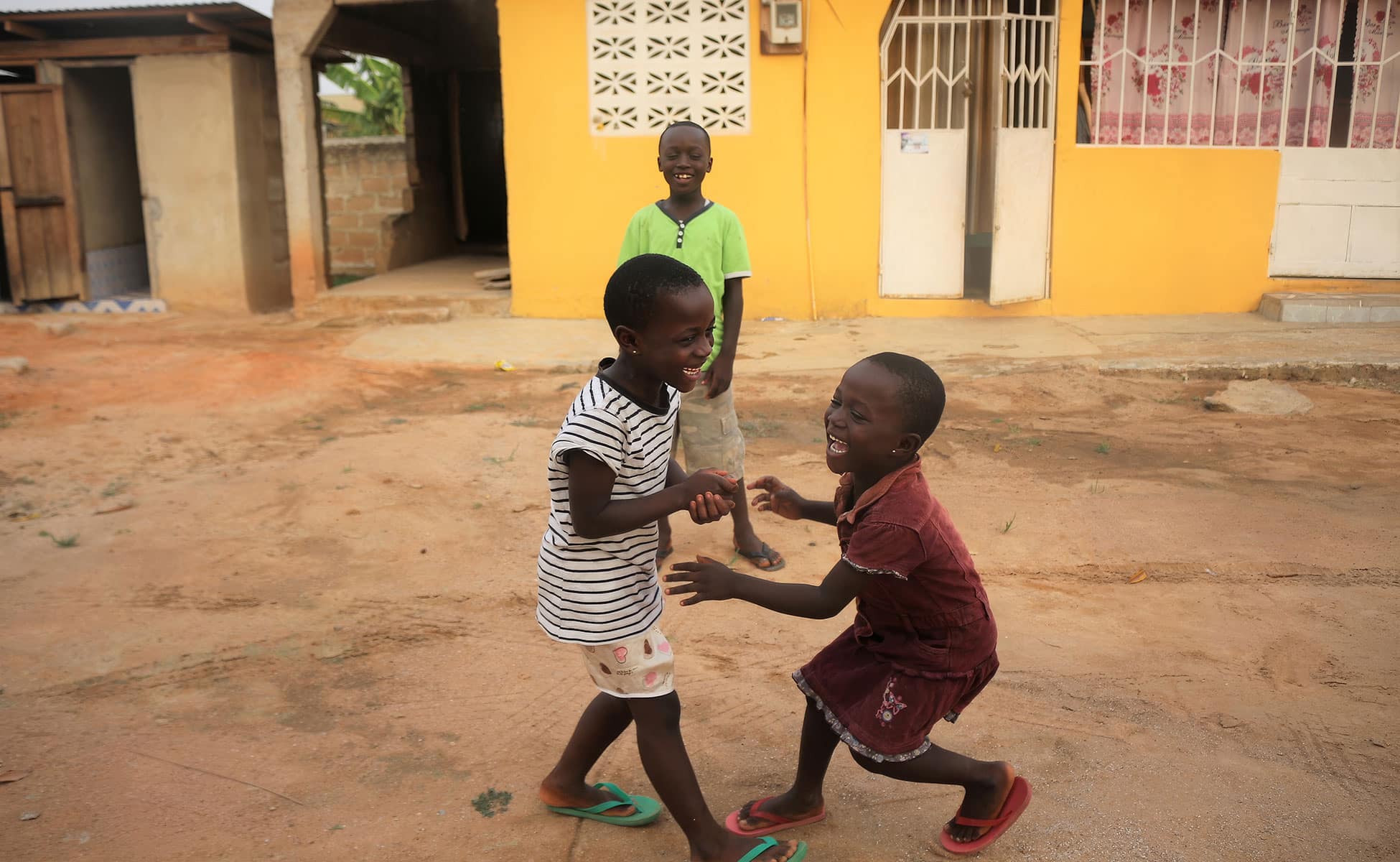 To barn fra Ghana, ei jente med kort hår, stripete t-skjorte, mønstrete shorts og grønne slippers, den andre med kort hår, rød kjole og røde slippers, ser mot hverandre og ler. I bakgrunnen står en gutt med grønn t-skjorte og kaki shorts. De står midt i ei gate med lys leire, i bakgrunnen en gul vegg. Foto: SOS-barnebyer