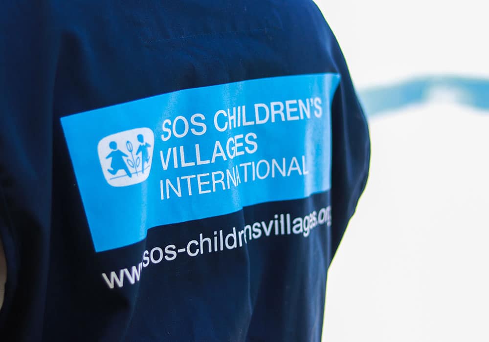 Ryggen  til en SOS-medarbeider i et nødhjelpsteam, som har på seg blå jakke med SOS-barnebyers internasjonale logo. Foto Lydia Mantler
