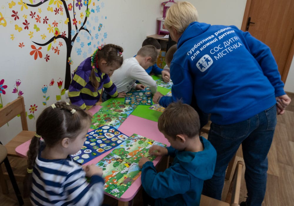 Flere barn sitter rundt et bord og tenger på og klipper i fargerikt papir. En medarbeider med lyst kort hår og blå genser med SOS-barnebyers logo på ryggen, hjelper dem. Foto: Katerina Ilievska