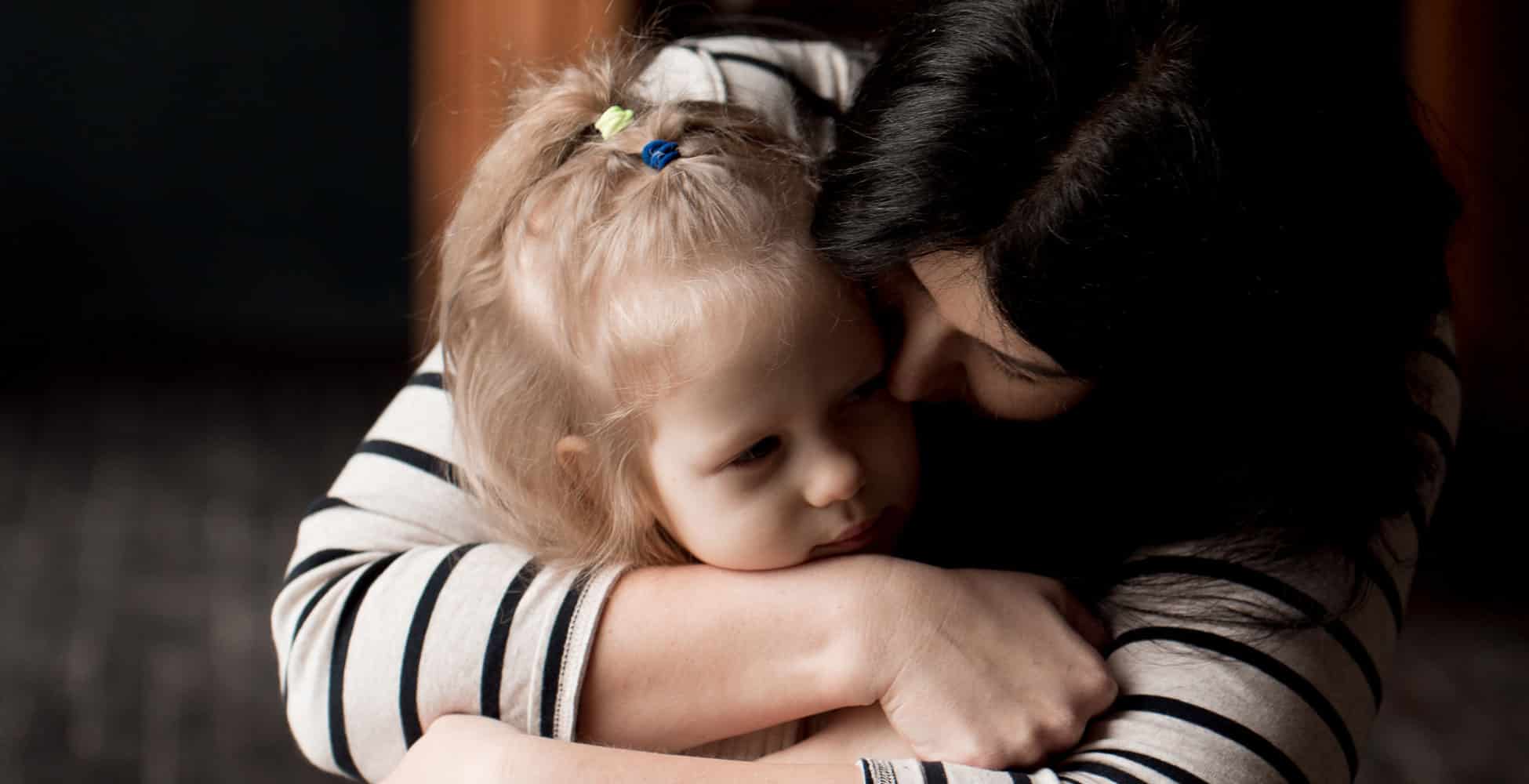 En mor med mørkt, langt hår og blå og hvit stripete genser ser ned og holder rundt den lille datteren sin, med lyst langt hår med spenner, hun sitter tullet inn i et pledd. Foto: Katerina Ptakha