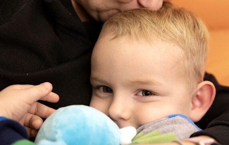 En mor med mørkt hår og mørk genser holder rundt den lille sønnen sin og kysser han på hodet. han har lyst kort hår, stripete blå, grå og grønn genser og holder en lyesblå bamse i armene. Foto: Katerina Ilievska