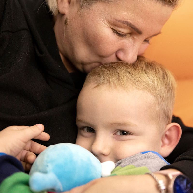 En mor med mørkt hår og mørk genser holder rundt den lille sønnen sin og kysser han på hodet. han har lyst kort hår, stripete blå, grå og grønn genser og holder en lyesblå bamse i armene. Foto: Katerina Ilievska