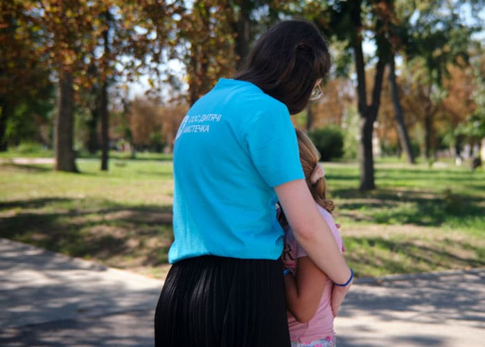 Ukraina: SOS-medarbeider med blå t-skjorte med SOS-barnebyers logo på ryggen holder rundt ei jente med lyst hår i hestehale, rosa strikk i håret og rosa t-skjorte. Illustrasjonsfoto Katerina Ilievska