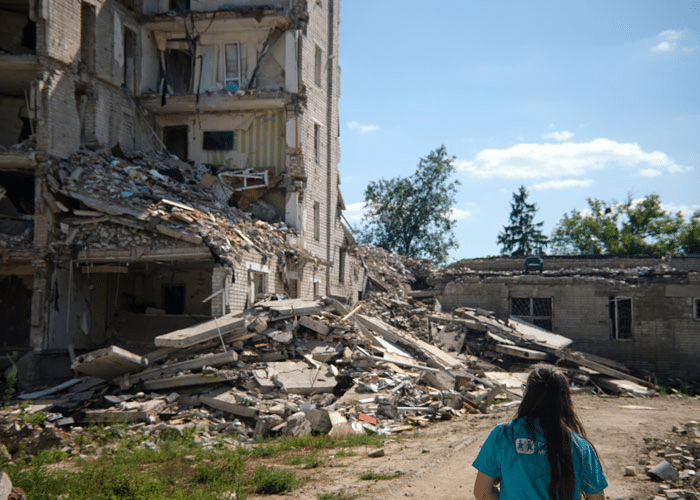 SOS-medarbeider med blå t-skjorte med SOS-barnebyers logo står med ryggen til og ser på ruinene av bygninger som er rammet. Foto: Katerina Ilievska