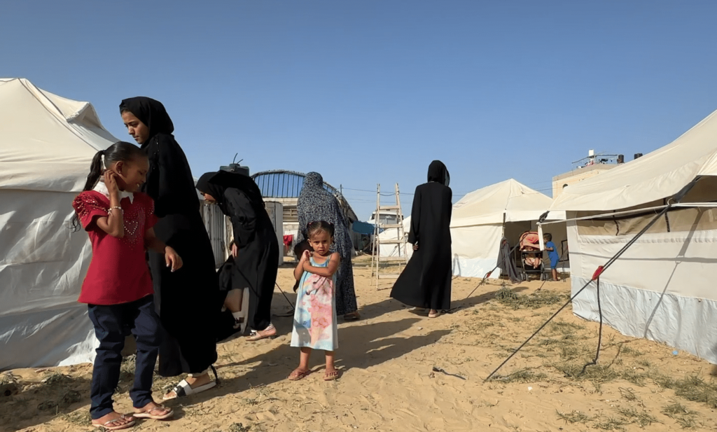 Liten jente i rosa og blå kjole som ser i kamera mellom telt i teltleir i Gaza