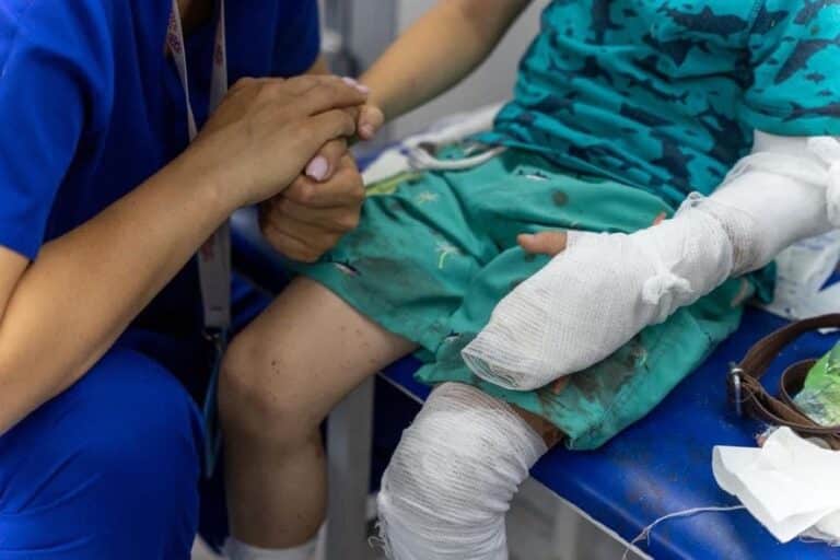 En pleier i blå uniform sitter på huk og holder hånden til et barn etter missilangrepet som traff barnesykehuset i Kyiv. Begge sitter og ansiktene er ikke med i bildet. Barnet har grønne skitne klær. Venstre arm og ben er bandasjert.
