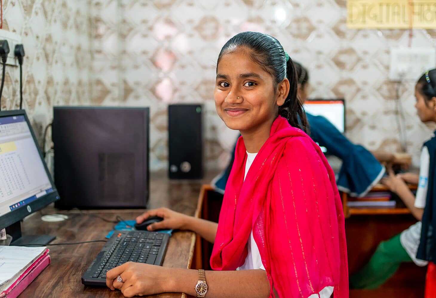 Nazia, med mørkt, langt hår i hestehale, rødt langt skjerf og hvit t-skjorte med SOS-barnebyers logo, sitter foran PCen og jobber. Det er flere barn og unge i rommet som også sitter foran PCer. Foto: Lydia Mantler