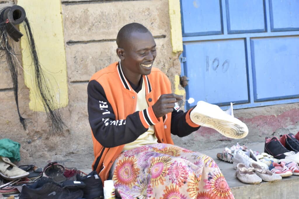 Anthony sitter på fortauskanten og reparerer sko. Han har på seg oransje jakke, gul t-skjorte og på fanget et mønstrete klede for å beskytte buksene. Foto: Saitoti Javason