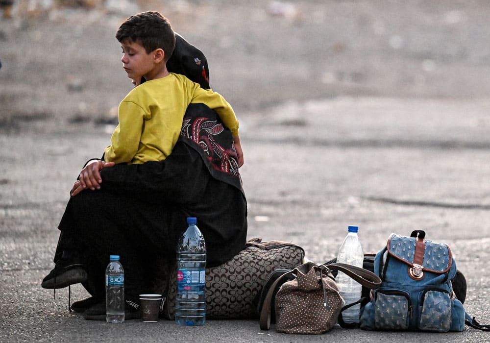 Mor med svare klær sitter på en veske med sønnen sin på fanget, han har mørkt, kort hår, gul genser og svart bukse. Ved siden av dem står det flere vannflasker og bager. Foto: Louai Beshara/AFP