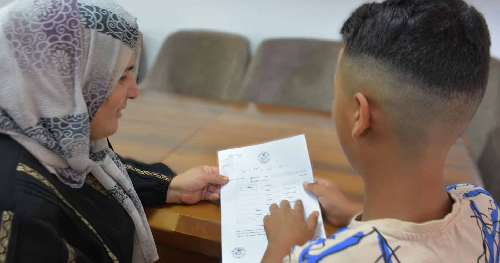 Gaza: Ammar, med kortklippet hår og mønstrete T-skjorte, sitter ved en pult og holder et ark med resultater fra en skoleprøve. Denne viser han til SOS-fostermoren sin, hun har mønstret hijab og svart kjole. Foto Lama Qattush