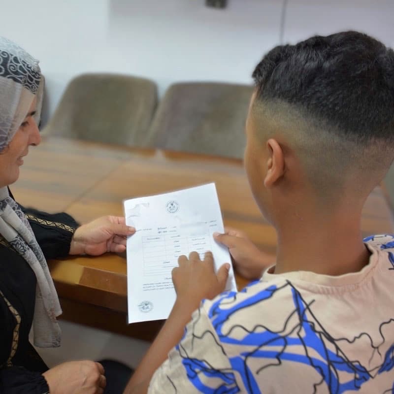 Ammar, med kortklippet hår og mønstrete T-skjorte, sitter ved en pult og holder et ark med resultater fra en skoleprøve. Denne viser han til SOS-fostermoren sin, hun har mønstret hijab og svart kjole. Foto Lama Qattush