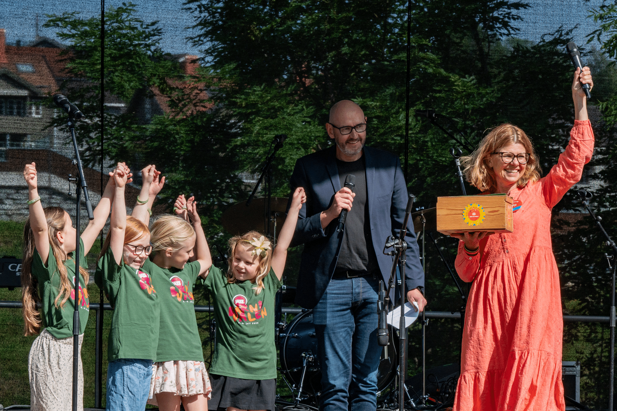 Bilde fra scenen i forbindelse med konserten Sangskattekista. Til venstre står flere barn med grønne t-skjorten og strekker hendene i været, i midten konferansieren med kort grått hår, skjegg, briller, blå dressjakke, svart t-skjorte og jeans,, og til høyre SOS-barnebyers frivillige leder i Kongsberg Kristina Schnell, hun har blondt halvlangt hår, briller og oransje kjole. I hånda holder hun ei trekiste med SOS-barnebyers logo. Foto: Birgit Fostervold