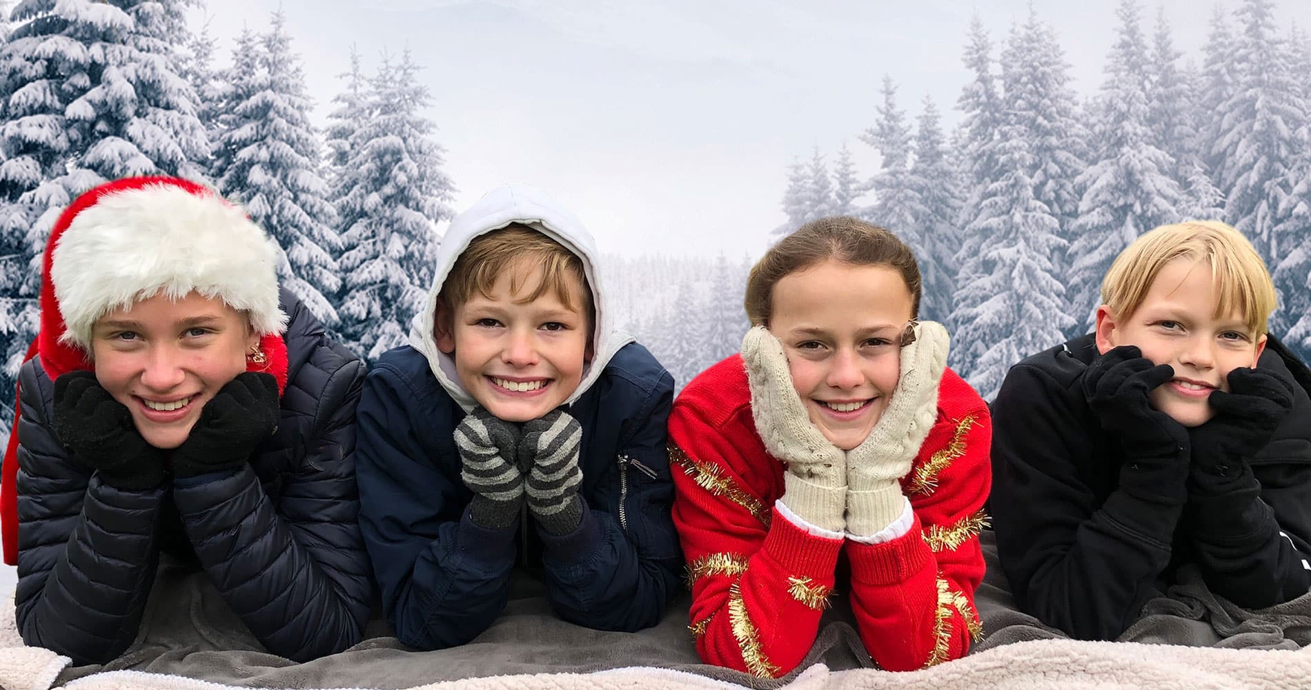 Fire barn ligger med hode i hendene og ser inn i kamera. Ei jente har nisselue, ei annen rød genser med glitterbånd. I bakgrunnen er det snøkledd skog. Foto SOS-barnebyer