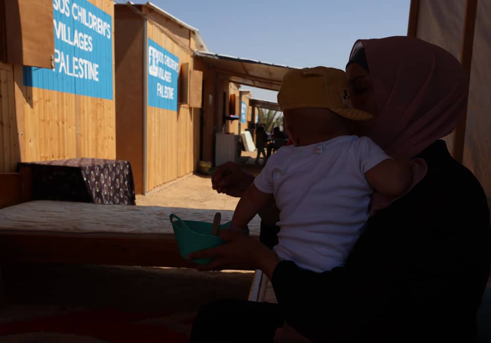 En mor med mørk hijab har den lille gutten sin på armen. I bakgrunnen ser vi SOS-barnebyers midlertidige leir, der husene er laget av planker. Foto Mohammad AlBaba