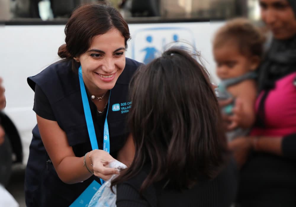 En av SOS-barnebyers medarbeidere i Syria, med mørkt langt hår og marineblå vest med SOS-barnebyers logo bøyer seg mot ei lita jente med mørkt krøllete, langt hår og deler ut en pose med mat og drikke. I bakgrunnen står moren, hun har et lite barn på armen. Foto SOS-barnebyer Libanon