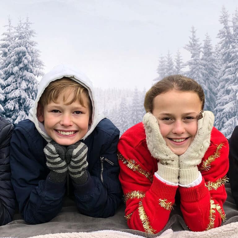 To barn ligger med hode i hendene og ser inn i kamera. Ei jente har nisselue, ei annen rød genser med glitterbånd. I bakgrunnen er det snøkledd skog. Foto SOS-barnebyer