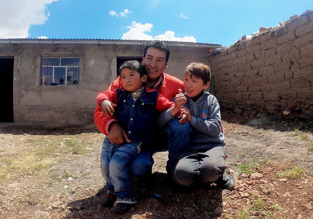 Fra pappaprogrammet i Peru. en far sitter på huk foran huset sitt, han har de to sønnene på fanget. Foto Julie Nilsen