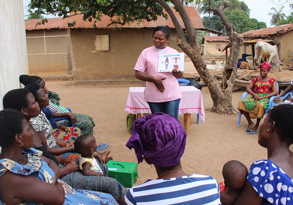 Togo: En av SOS-barnebyers medarbeidere, med rosa t-skjorte og jeans, står foran en gruppe kvinner  som sitter ute blant hus lagt av leier og stein. Foto Pierre Devé