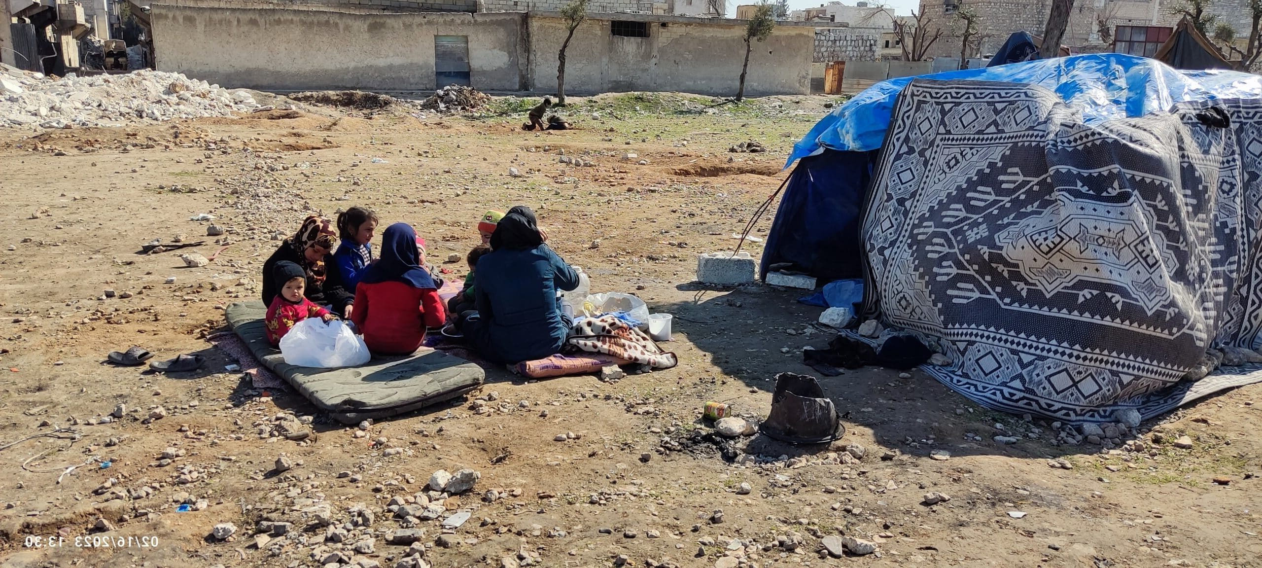 En familie i Aleppo sitter på et teppe og en madrass utendørs, omgitt stein, rusk og jord I forgrunnen ser vi et telt dekket av et mønstret stoff og blå plast, sannsynligvis for å beskytte mot været. Familien med flere barn, sitter i en sirkel. De sitter i et flyktnings- og nødhjelpsområde I bakgrunnen er det ødelagte bygninger.