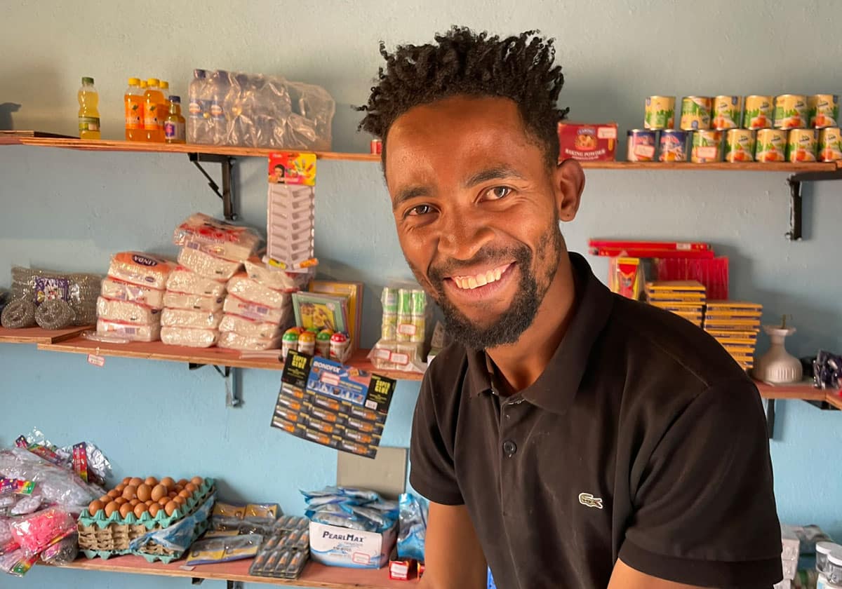 Antonio står inne i butikken. Han har krøllet svart hår, svart t-skjorte med krage, hodetelefoner rundt halsen og smiler. Det er mange forskjellige varer i hyllene på veggen. Foto Turid Weisser