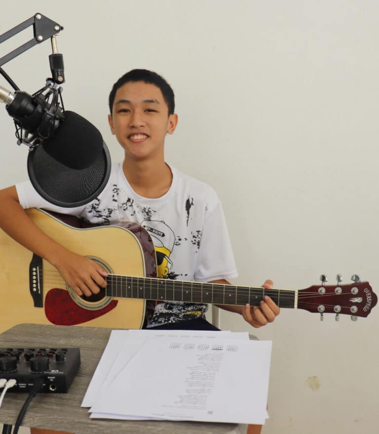 Gutt fra et Thailand med mørkt, kort hår og hvit genser, sitter ved et bord og spiller gitar. Han tar opp det han synger og spiller og har en mikrofon og opptaksutstyr. Foto: SOS-barnebyer