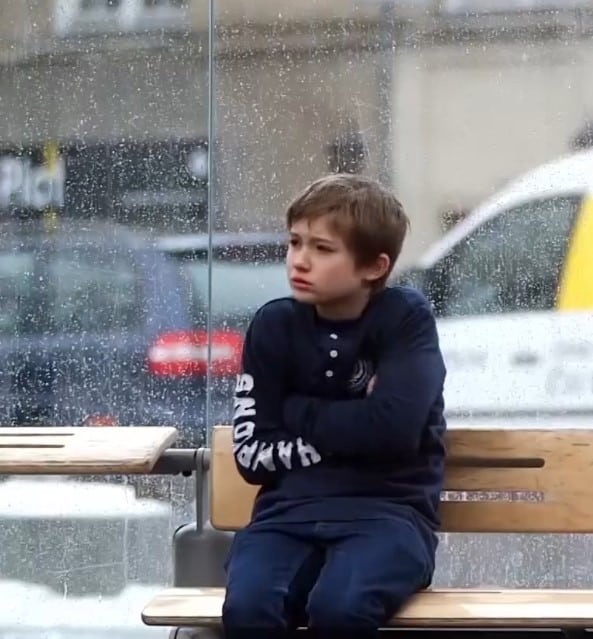 En gutt med mørkt, blondt hår, blå genser og jeans sitter på en bussholdeplass uten jakke og fryser, det er snø i lufta. Foto: fra filmen - SOS-barnebyer