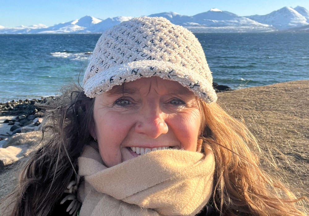 Elisabeth Kvinnesland, med langt, bølgete, blondt hår, hvit strikkelue og lyst skjerf er fadder i SOS-barnebyer. Hun står på ei strand, i bakgrunnen ser vi havet og høye snødekte fjell. Foto Privat