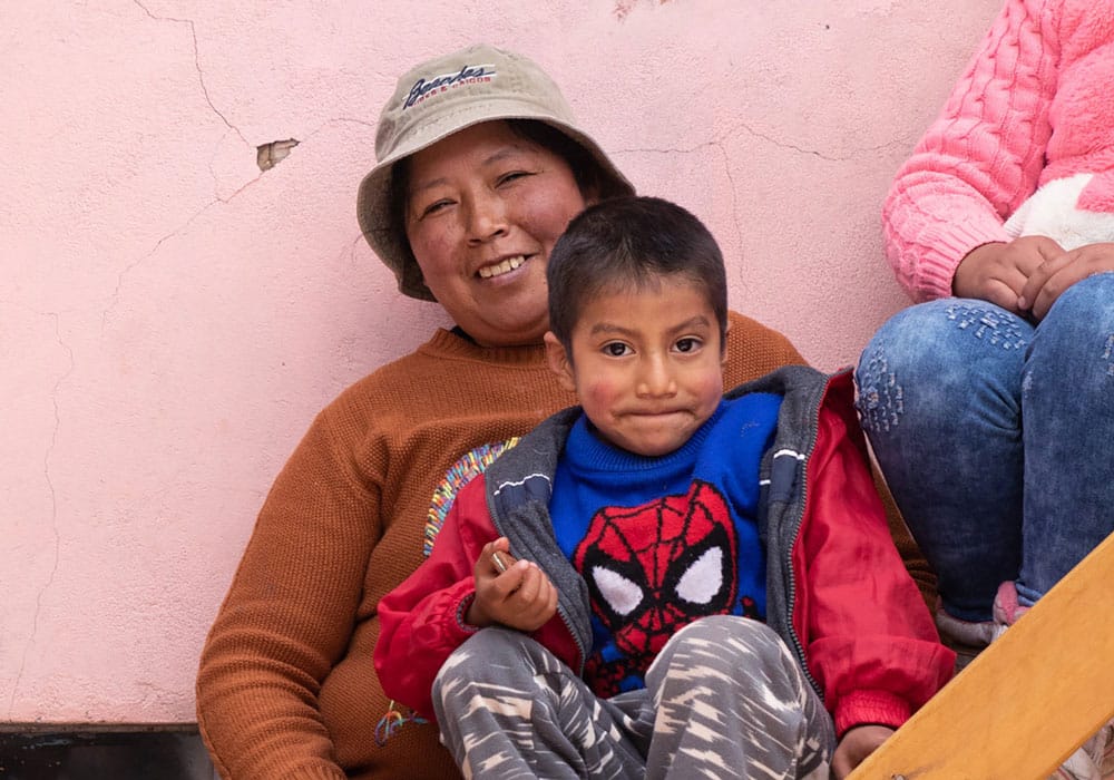 En mor fra Peru med beige bøttehatt og brun genser sitter i ei trapp med sønnen på fanget. Han har spidermangenser, rød jakke og mønstrete bukse og smiler lurt. Datteren sitter øverst i trappa, hun har hvit lue, rosa hettejakke og jeans. Bak dem er det en rosa murvegg. Foto Alejandra Kaiser