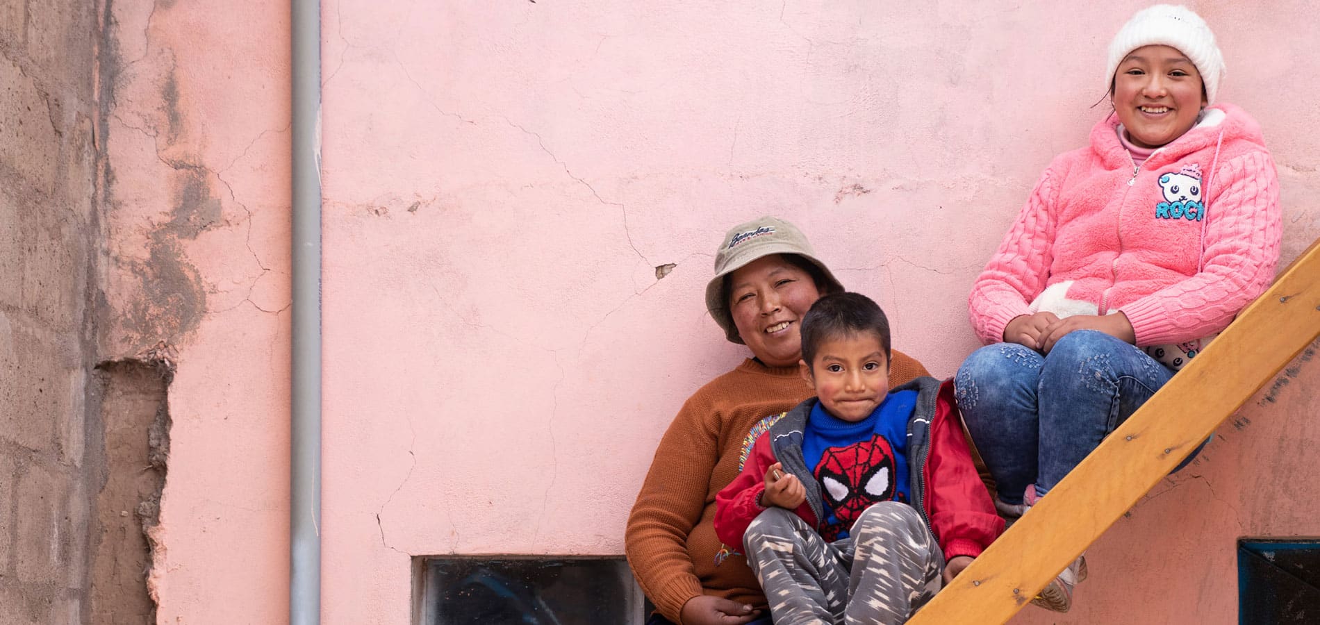 Vilje til enring: En mor fra Peru med beige bøttehatt og brun genser sitter i ei trapp med sønnen på fanget. Han har spidermangenser, rød jakke og mønstrete bukse og smiler lurt. Datteren sitter øverst i trappa, hun har hvit lue, rosa hettejakke og jeans. Bak dem er det en rosa murvegg. Foto Alejandra Kaiser