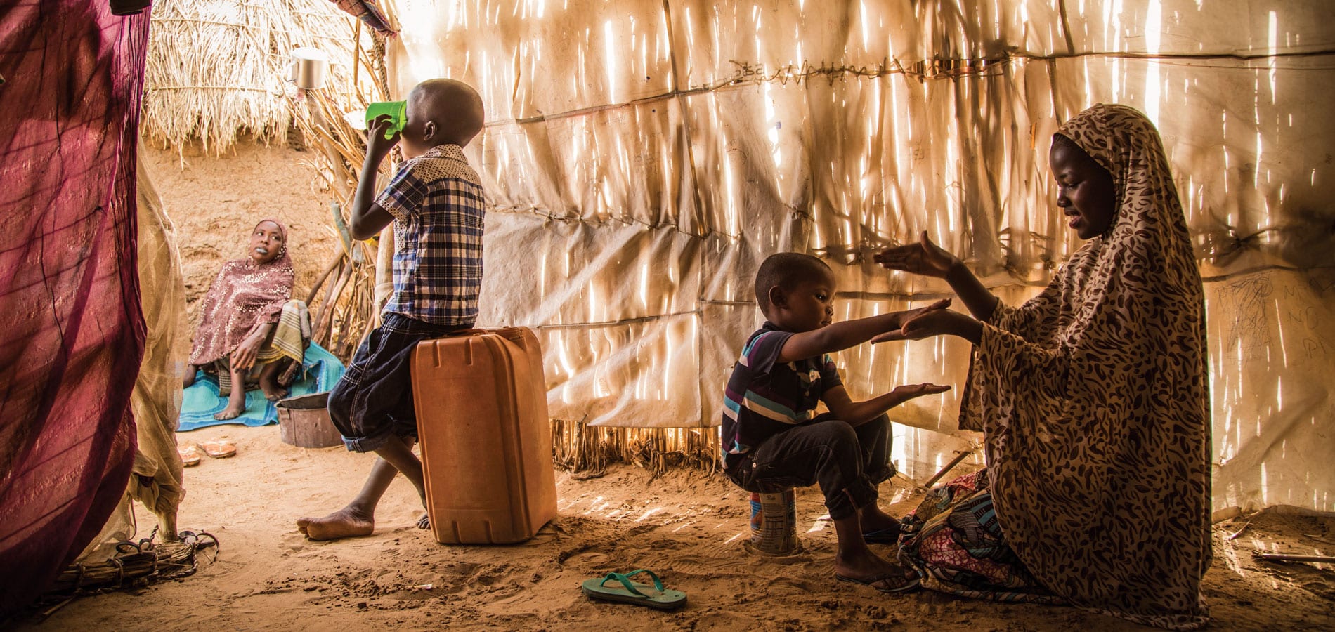 En mor fra Niger og barna hennes sitter inne i ei hytte laget av strå. Hun leker med det minste barnet, en av sønnene sitter på ei plastkanne og drikker av en grønn kopp. Foto Vincent Tremeau