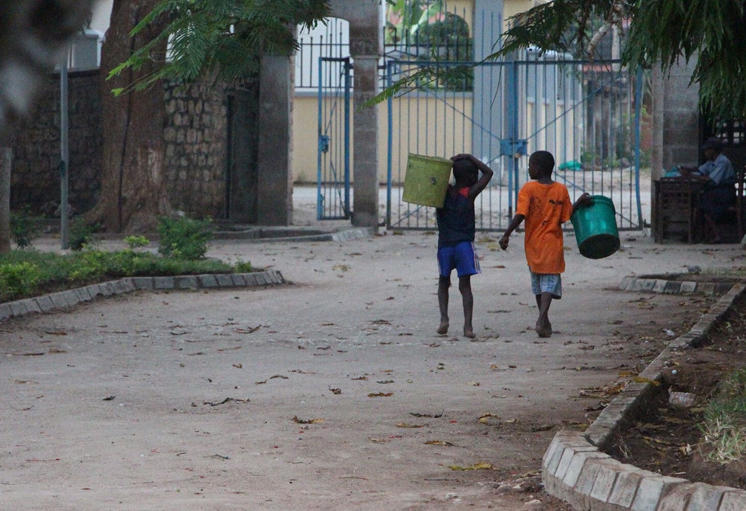 To gutter, begge med shorts og t-skjorte, bærer store plastikkspann går å en vei. i bakgrunnen er det et høy port. Illustrasjonsfoto fra Tanzania Line Grove Hermansen