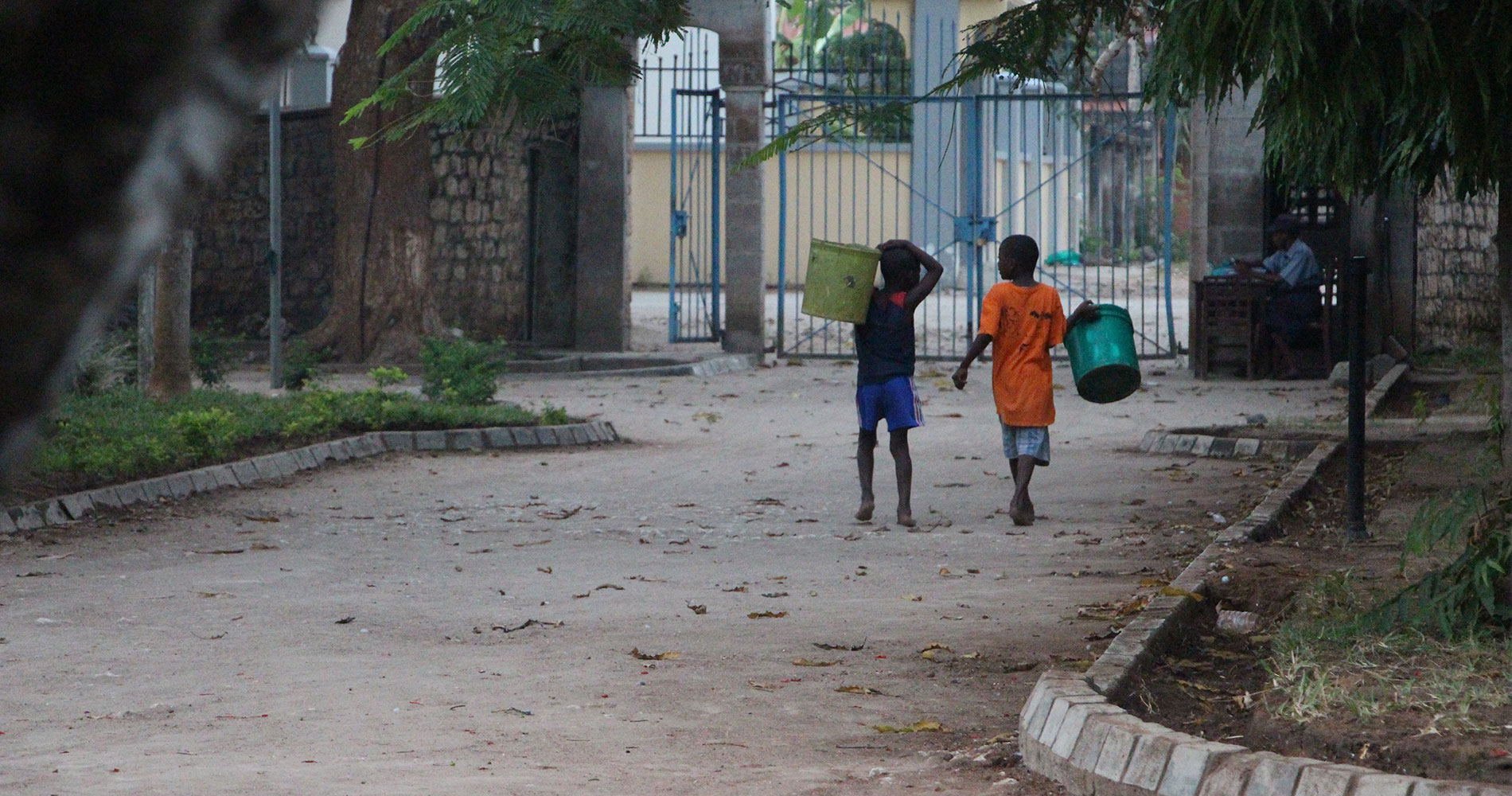 To gutter, begge med shorts og t-skjorte, bærer store plastikkspann går å en vei. i bakgrunnen er det et høy port. Illustrasjonsfoto fra Tanzania Line Grove Hermansen