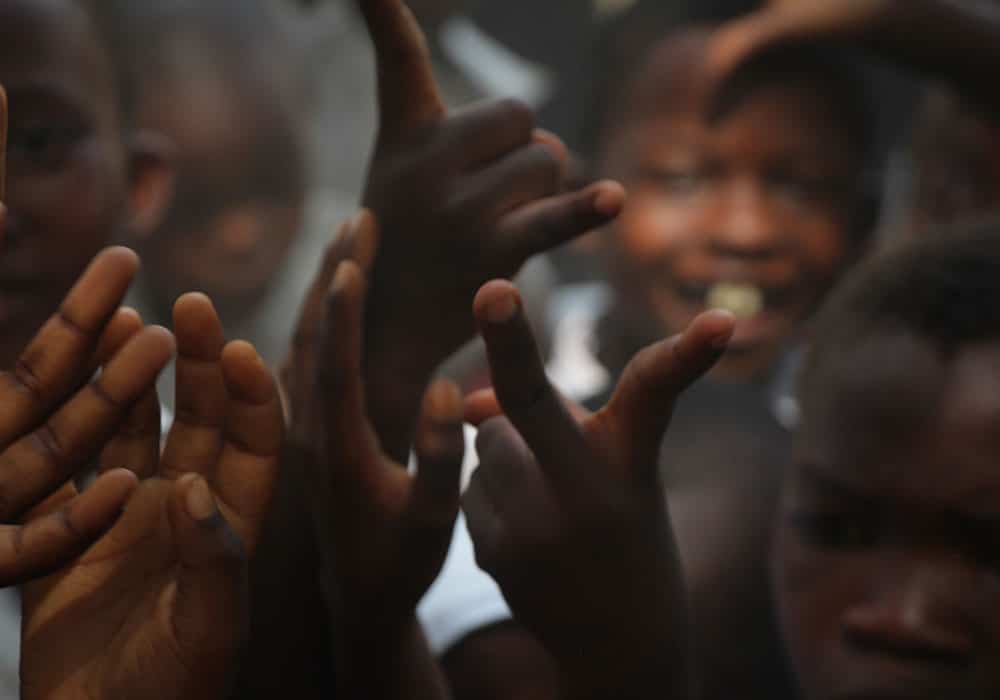 Mange barn fra Tanzania streker hendene i været, ansiktet til et barn som smiler skimtes i bakgrunnen. Foto: Line Grove Hermansen