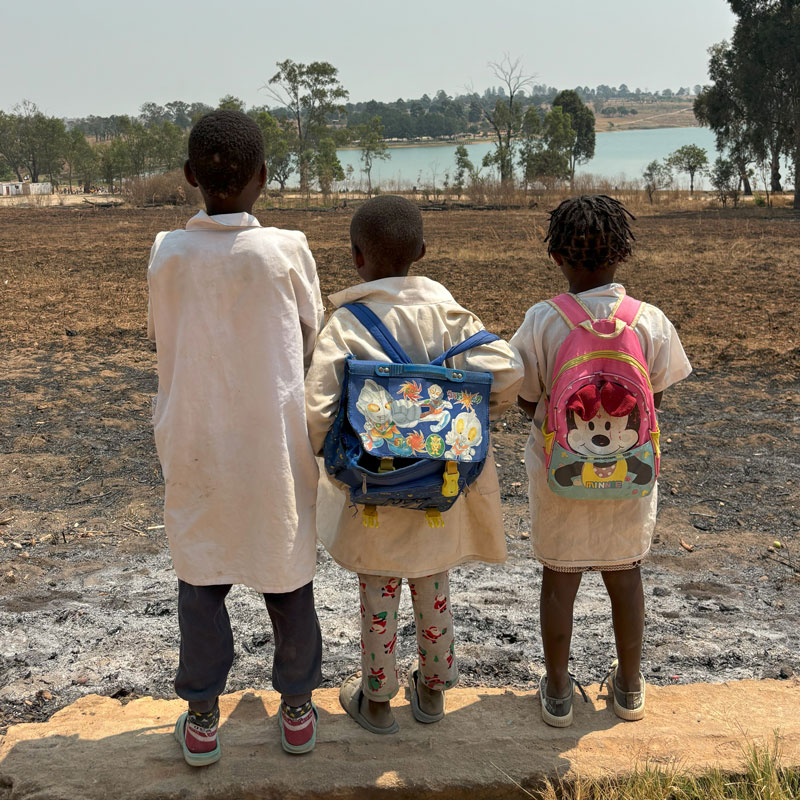 Tre barn som får støtte gjennom Fagforbundets engasjement i Huambo, står på en murkant med ryggen til og ser utover et jorde og en innsjø. alle tre har skolefrakker, to av dem har sekk på ryggen med flotte motiver. Foto Sigurd Skjefstad