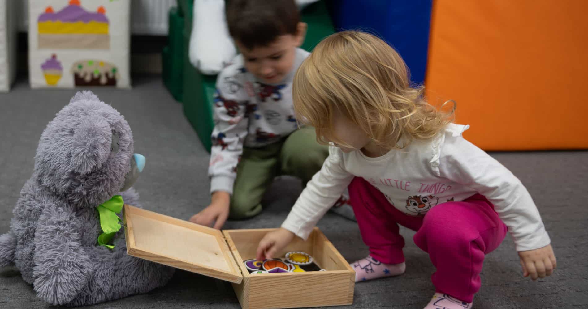 Ukraina: Trygge steder for lek er med på å bearbeide traumer. Ei lite jente med lys halvlangt hår, hvit genser og rosa bukser sitter på huk og leker med noen klosser. En bamse sitter på gulvet og en liten gutt sitter i bakgrunnen. Foto Katerina Ilievska