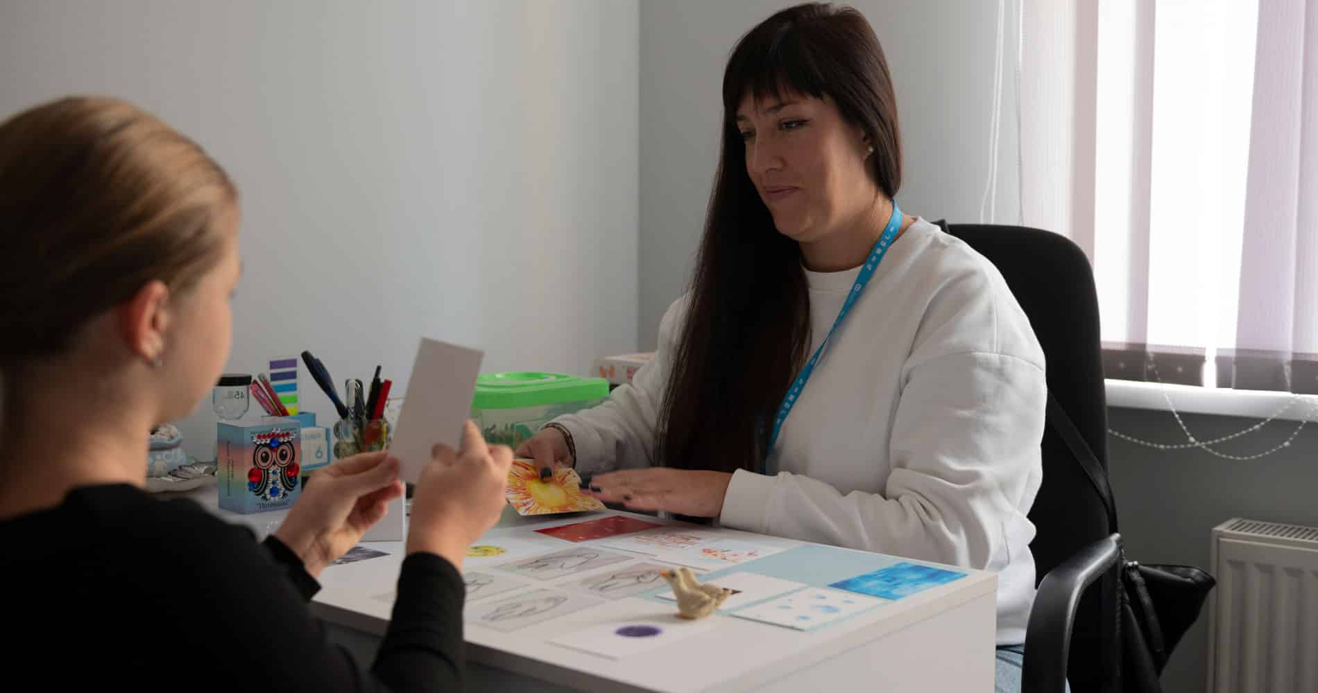 Iryna, psykolog i SOS-barnebyer Ukraina. med langt mørkt hår og hvit genser, sitter ved et bord sammen med ei jente. Hun har blondt hår og mørk genser. Foto Katerina Ilievska