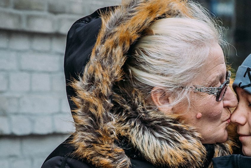 Bestemoren med grått hår i topp, briller og vinterjakke med pelskrage kysser et av barnebarna, Han er anonymisert og vi ser bare litt av nese og kinn. Foto: Alea Horst
